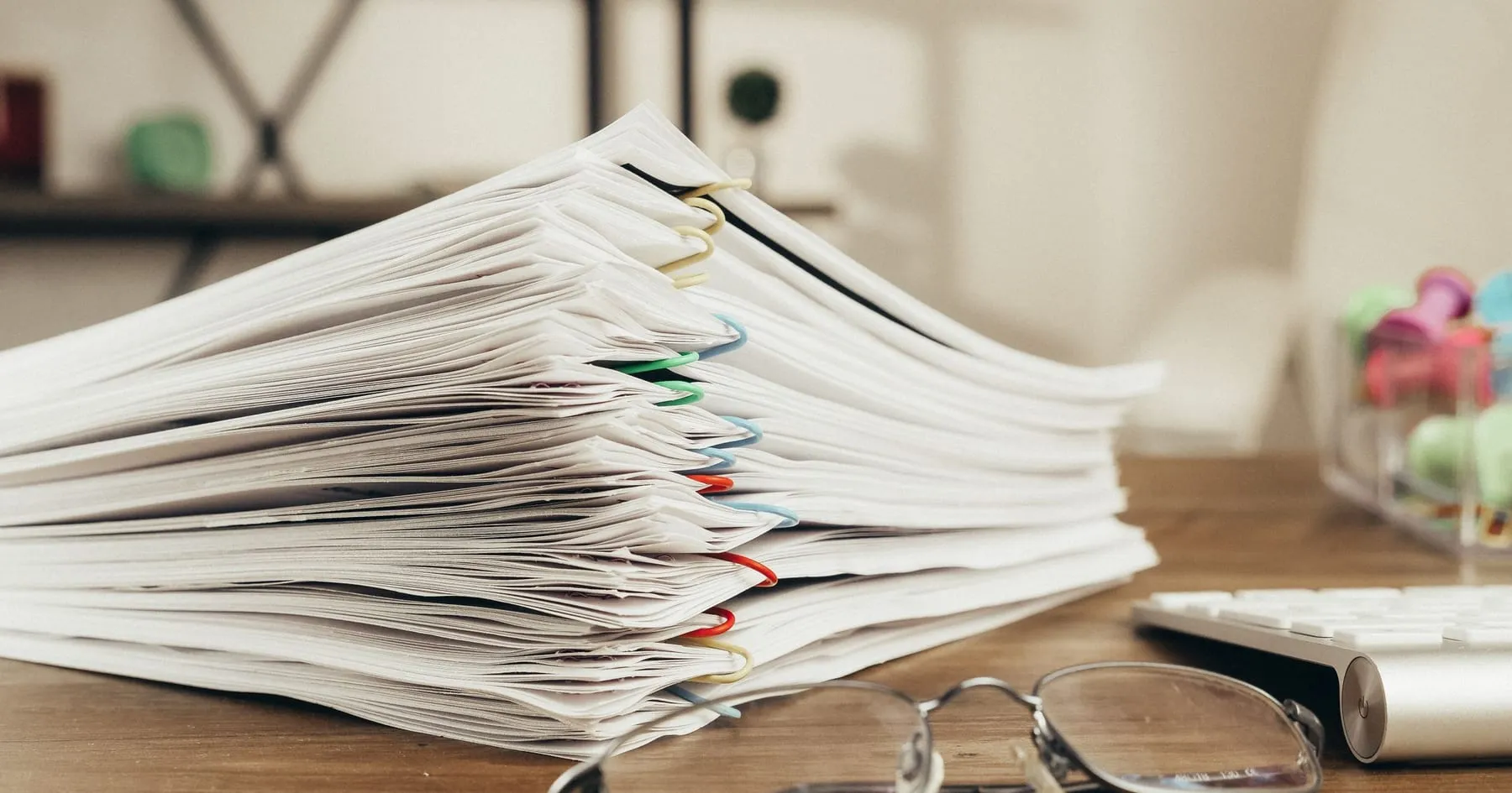 Haute pile de documents administratifs retenus par des trombones de couleur, posée sur un bureau en bois à côté d'une paire de lunettes et d'un clavier.